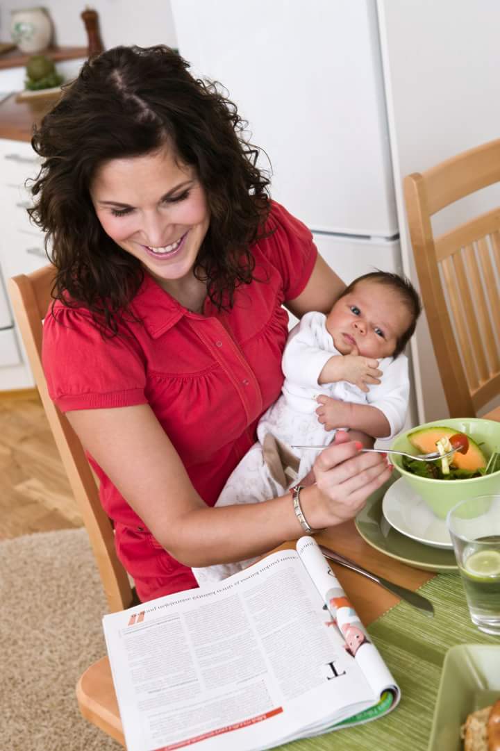 Äiti lukee lehteä ruokapöydässä, vauva sylissä. 
